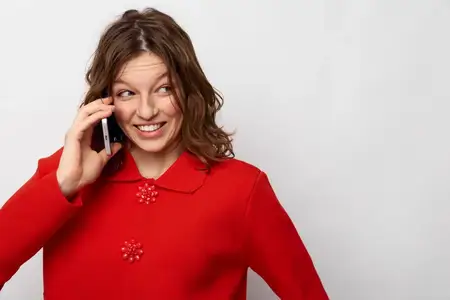 Счастливая молодая женщина в красном трикотаже держит телефон у уха, улыбаясь и глядя в сторону