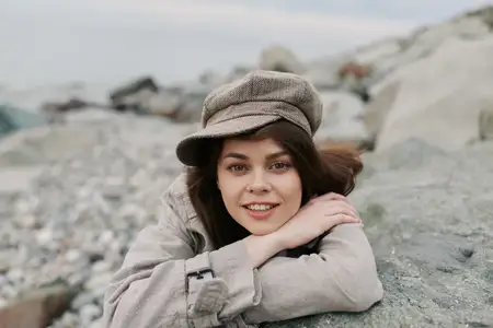 Улыбающаяся молодая женщина в клетчатой кепке и пальто отдыхает на каменистом берегу на фоне природы
