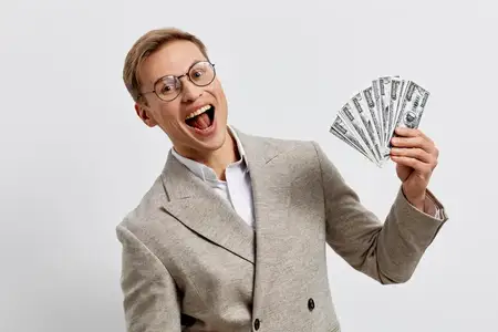 Взволнованный молодой европеец в бежевом клетчатом пиджаке держит долларовые купюры, улыбаясь и празднуя финансовый успех, фриланс-доход, онлайн-работа, образ жизни цифрового кочевника
