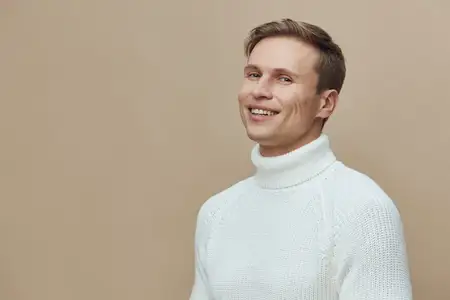 Жизнерадостный мужчина в белом свитере улыбается и смотрит в камеру, стоя на простом бежевом фоне