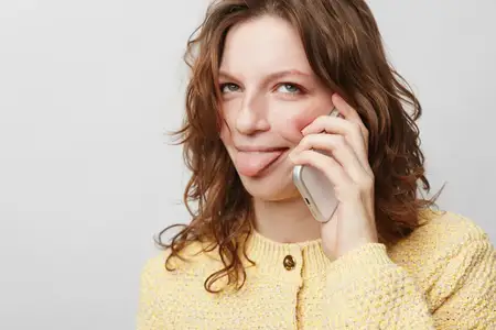 Улыбающаяся женщина в желтом вязаном свитере прижимает телефон к уху и игриво показывает язык