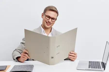 Улыбающийся мужчина работает с папкой в современном офисе, он в очках и повседневном пиджаке, сидит за столом с ноутбуком и блокнотом, изолирован на белом фоне