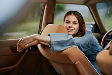 Счастливая молодая женщина в джинсовой куртке, облокотившаяся на винтажный автомобиль, демонстрирует беззаботный дух и радость в яркой и солнечной обстановке, что идеально подходит для тем…
