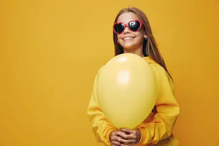 Юная девушка в солнцезащитных очках в форме сердца и желтом свитере держит воздушный шар с жизнерадостной улыбкой и ярким стилем на сплошном желтом цветном фоне