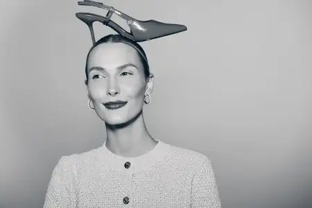 Белая женщина со светлой кожей и серьгами позирует на однотонном фоне, удерживая на голове туфлю на высоком каблуке