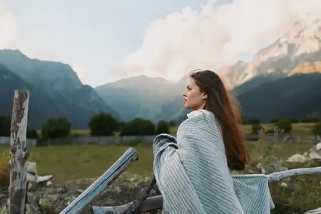 Женщина, укутанная в уютное одеяло, стоит у деревянного забора, смотрит на горный пейзаж, наслаждается спокойным образом жизни на открытом воздухе на фоне природного окружения