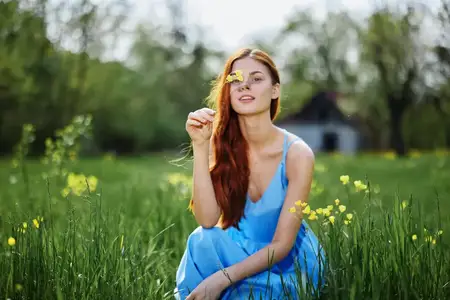 Женщина весной на природе улыбается, обнажая зубы, сидя в поле на зелёной траве среди цветов, и улыбается счастливому закату в синем платье с рыжими волосами