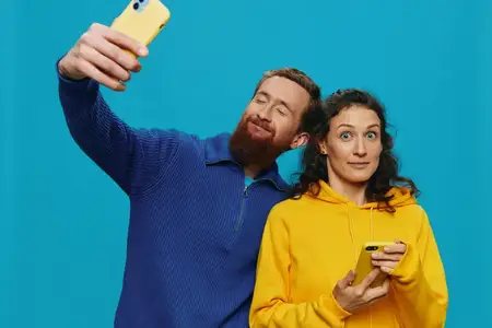 Забавная пара — женщина и мужчина с телефонами в руках, делающие селфи с кривыми улыбками на синем фоне