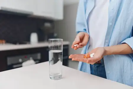 Женщина принимает лекарство, запивая его водой из стакана; на кухонной стойке стоит пузырёк с таблетками