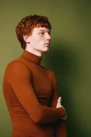 Портрет рыжеволосого молодого человека в коричневой водолазке с длинным рукавом, позирующего вполоборота с нейтральным выражением лица на фоне сплошного зеленого фона в студии