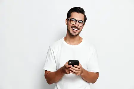 Счастливый улыбающийся мужчина в очках держит смартфон, стоя перед однотонным белым фоном