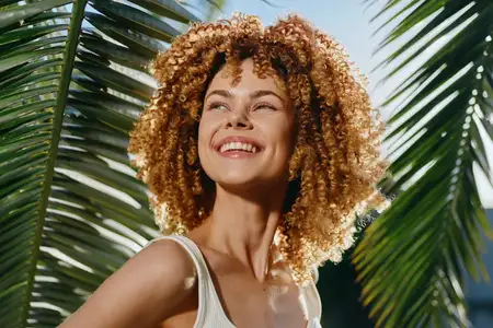Улыбающаяся женщина с кудрявыми волосами стоит на фоне ярких пальмовых листьев, наслаждаясь теплым солнечным светом и излучая радостные, беззаботные летние вибрации