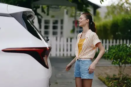 Веселая молодая женщина стоит рядом со своим белым автомобилем, одетая в полосатую рубашку и джинсовые шорты