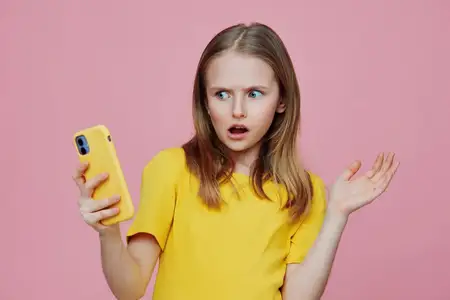 Удивленная девочка в желтой рубашке держит телефон на розовом фоне