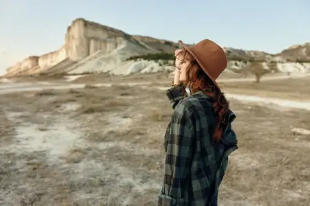 Женщина, любительница приключений на открытом воздухе, в клетчатой рубашке и шляпе стоит перед величественным горным хребтом