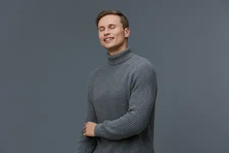 Улыбающийся мужчина в сером вязаном свитере с водолазкой с закрытыми глазами наслаждается моментом цифрового детокса, чувствует себя расслабленным и умиротворенным, изолирован на однотонном сером…
