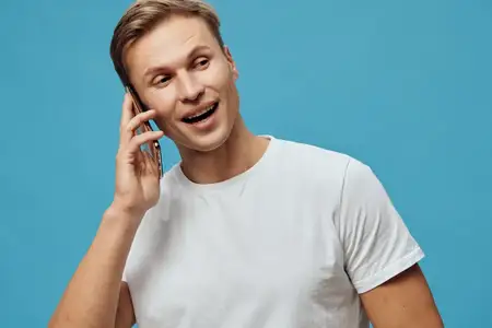 Счастливый мужчина с короткими волосами в повседневной белой футболке улыбается и разговаривает по мобильному телефону, изолирован на ярком синем фоне