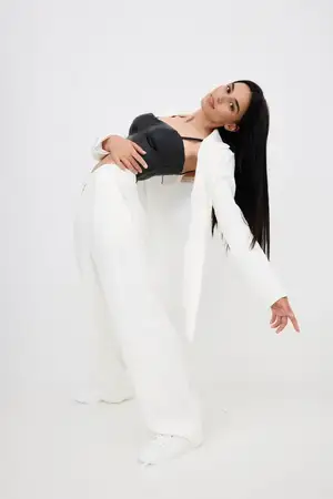 Женщина в белых широких брюках и чёрном топе без рукавов грациозно откидывается назад на фоне однотонного белого фона, демонстрируя современный стиль и элегантность