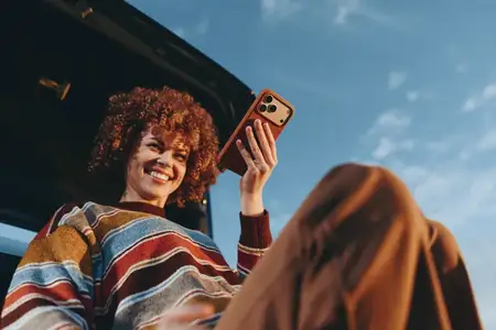 Улыбающаяся женщина с кудрявыми волосами держит смартфон для селфи, вид из салона машины, сцена на улице с небом и облаками, жизненный стиль путешествия, дружелюбный молодёжный портрет, дневной…