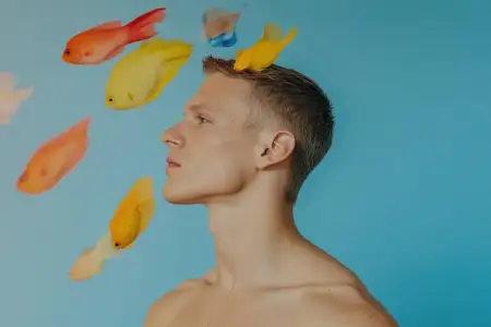 Юный мужчина-модель с задумчивым выражением лица изображен на ярком синем фоне в окружении разноцветных рыб, что символизирует творчество и вдохновение, выражая безмятежность
