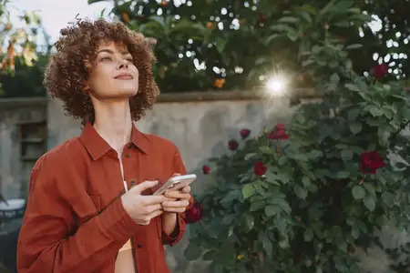 Лайфстайл-сцена с женщиной в саду, улыбающейся своему устройству, непринужденная поза, залитая солнцем зелень, интимная уличная атмосфера, естественные тона, современное настроение благополучия