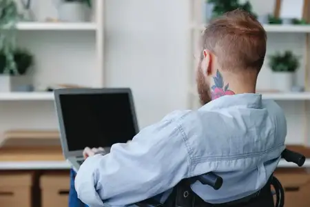 Мужчина в инвалидной коляске, фрилансер, работает за ноутбуком дома, вид со спины