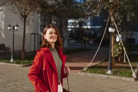 Стильная молодая женщина в красном пальто и зеленом свитере стоит на вымощенной площади, залитые солнцем деревья позади, она улыбается, глядя в камеру, создавая яркий модный момент в городе