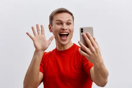 Веселый молодой мужчина-европеоид совершает видеозвонок, используя смартфон, в красной футболке, улыбается, делает возбужденный жест, машет рукой, изолирован на сером фоне