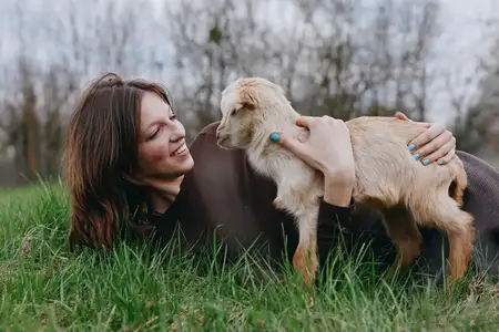 Молодая женщина с длинными каштановыми волосами лежит на зелёной траве в коричневом свитере, нежно обнимая козлёнка и улыбаясь