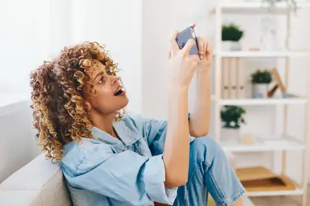Улыбающаяся женщина держит смартфон на уютном диване в гостиной, наслаждаясь онлайн-досугом