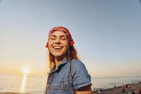 Улыбающаяся женщина, одетая в современный бохо-вестерн деним стиль и красную бандану, стоит на пляжной улице на закате