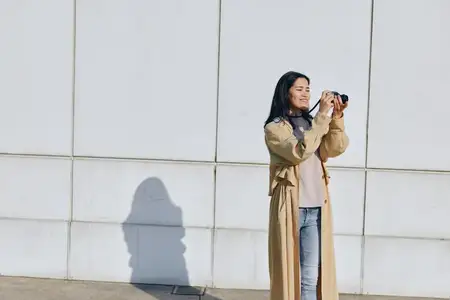 Сцена уличной фотографии: женщина использует камеру, одета в длинное тренч-пальто, на фоне простой стены, с акцентом на композицию, фокус и естественную городскую студийную атмосферу