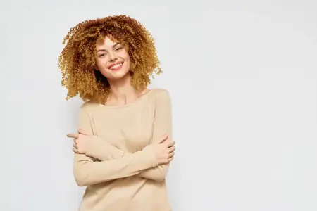 Улыбающаяся молодая женщина с вьющимися волосами, одетая в бежевый свитер, скрестила руки на груди на фоне светло-серого фона