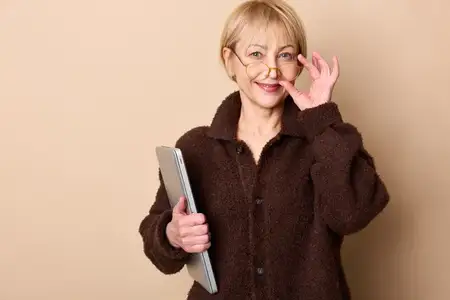 Пожилая женщина в очках мягко улыбается и машет рукой, держа планшет при студийном освещении
