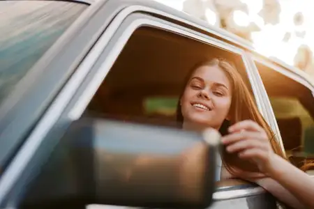 Радостная молодая женщина с длинными волосами выглядывает из окна машины, излучая счастье и волнение, наслаждаясь летним путешествием, полным приключений и свободы