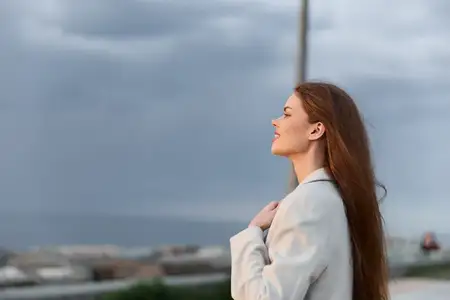 Женщина с длинными рыжими волосами в легком пальто стоит на улице под облачным небом, выражая спокойствие и надежду