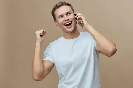 Взволнованный молодой светлокожий мужчина со смартфоном разговаривает по телефону, делая жест сжатого кулака, в повседневной белой футболке, изолирован на бежевом фоне, счастливые положительные…