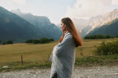 Женщина, закутанная в серое одеяло, стоит на гравийной дорожке на фоне гор и зелёных полей, отражая спокойный образ жизни и момент на природе