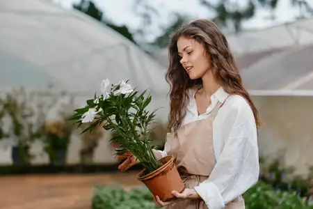 Веселая молодая женщина, одетая в мягкие тона, нежно держит в руках горшечный цветок в теплице, демонстрируя свою страсть к садоводству, природе и сезонной красоте