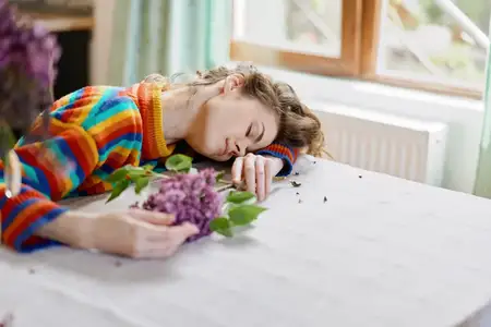 Спокойный домашний момент: женщина спит за столом с цветами, мягкий свет, непринужденная поза, домашний уют, нежные цвета, умиротворенное настроение и тихое творчество