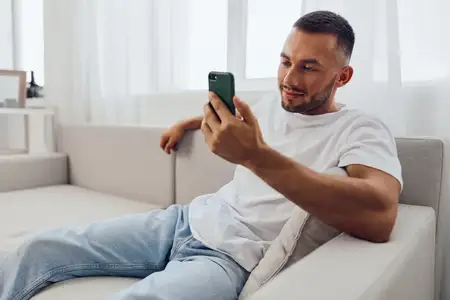 Веселый мужчина непринужденно листает свой смартфон, сидя на уютном диване