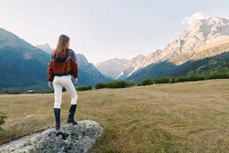 Женщина в походных ботинках осматривает травянистый луг и альпийскую тропу, любуясь впечатляющими горами и ясным небом
