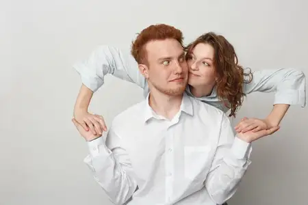 Игривые молодые мужчина и женщина с рыжими и вьющимися волосами в белых рубашках улыбаются и тесно взаимодействуют
