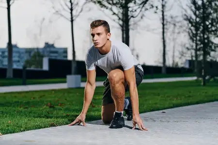 Спортивный мужчина в шортах и футболке готовится к пробежке на природе, в парке, возле деревьев, на улице