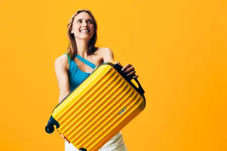 Женщина уверенно стоит с жёлтым чемоданом на оранжевом фоне, излучая яркость и смелость