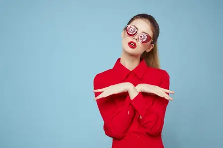 Женщина в красной рубашке на синем фоне держит руки у лица, в солнцезащитных очках, с красными губами, макияж, модель