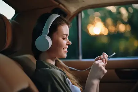 Улыбающаяся молодая женщина в наушниках слушает музыку на смартфоне, сидя в автомобиле на фоне красивого заката, выражая радость и расслабление
