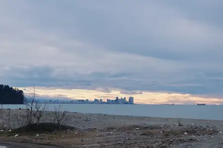 Прибрежная сцена с водой и городским пейзажем вдалеке, свечение на горизонте, облачное небо и пересеченная местность на переднем плане, умиротворенное настроение, подходящее для тем путешествий…