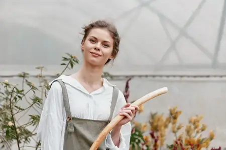 Счастливая молодая женщина в повседневной садовой одежде держит деревянный садовый инструмент в теплице на фоне красочных растений, демонстрируя любовь к природе и садоводству
