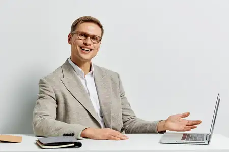 Улыбающийся мужчина работает за ноутбуком в повседневной куртке и очках, сидя за белым столом с блокнотом и ручкой, изолированный на белом фоне